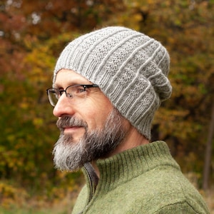 May include: A man wearing a gray knitted beanie and a green sweater. He is wearing glasses and has a beard.
