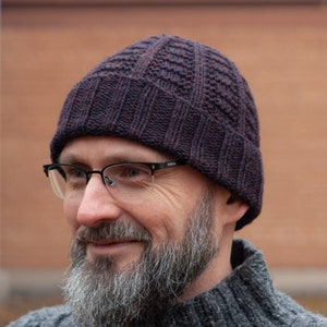 Pode incluir: Um homem com barba grisalha está usando um gorro de tricô roxo escuro com um padrão texturizado e óculos de armação preta. Ele está usando um suéter de tricô cinza com tranças.