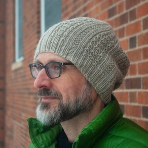 Pode incluir: Um homem usando um gorro de malha cinza com um padrão texturizado. Ele está usando uma jaqueta verde e tem barba.