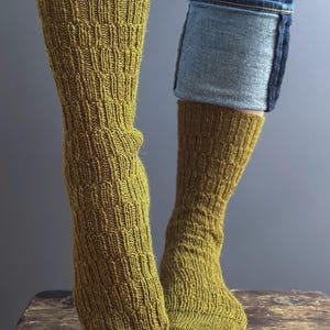 May include: Close-up of two legs wearing olive green knitted socks. The socks have a textured, patterned design and are paired with blue jeans. The socks are pulled up to the mid-calf.