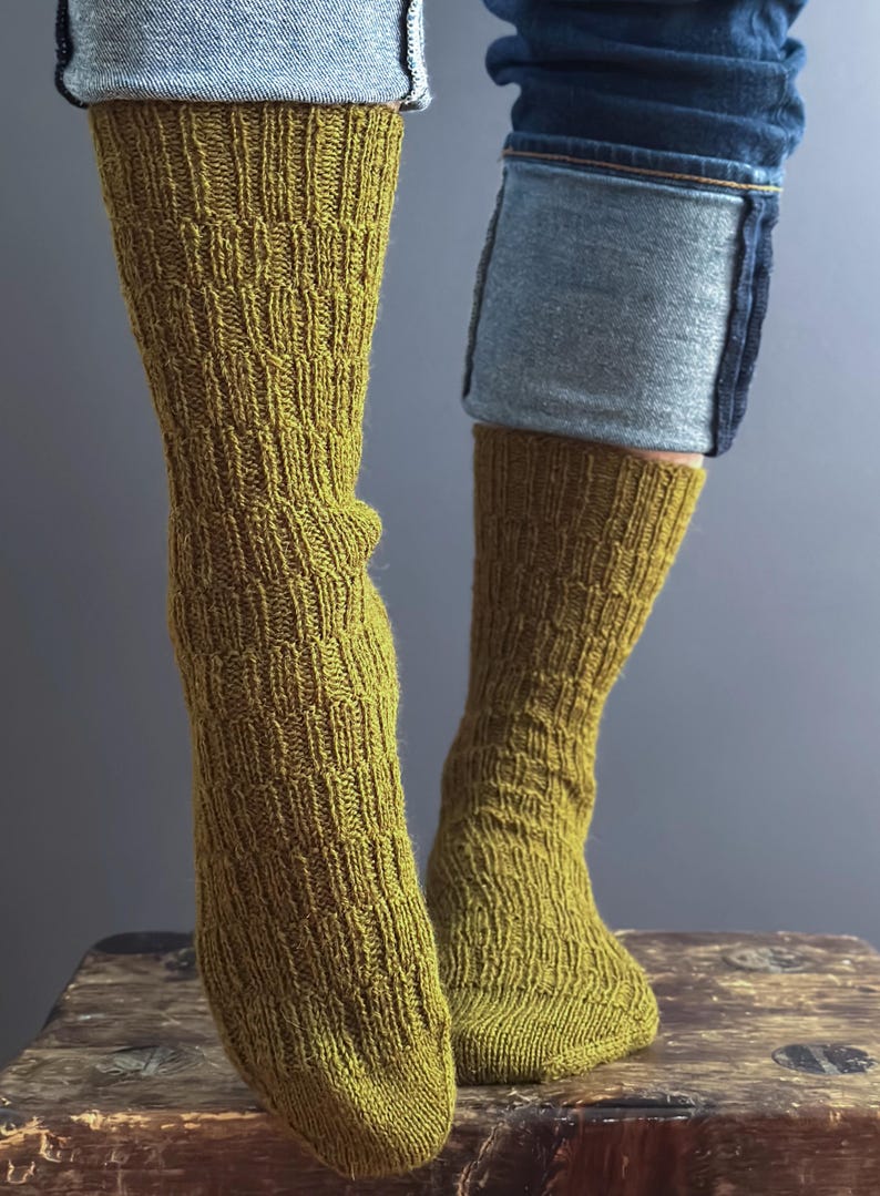 May include: Close-up of two legs wearing olive green knitted socks. The socks have a textured, patterned design and are paired with blue jeans. The socks are pulled up to the mid-calf.