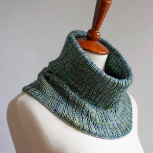 May include: A hand-knitted cowl in shades of blue, green, and yellow, displayed on a mannequin. The cowl features a textured, ribbed design and is made of soft yarn. This neck warmer is a cozy accessory for cold weather.