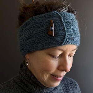 May include: A teal knitted headband with a wooden button closure. The headband is made of a thick, textured yarn. The wooden button is a dark brown color. The person in the image is wearing a gray turtleneck sweater.