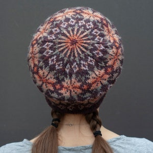 May include: A knitted hat with a colorful geometric pattern in shades of brown, pink, and green. The hat is worn by a person with brown hair pulled back in two braids.
