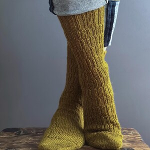 May include: Mustard yellow knitted leg warmers with a textured pattern. The leg warmers are worn with denim shorts. The knitted socks are made of a soft, warm material.