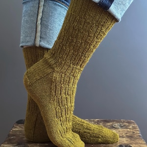 May include: A pair of olive green knitted socks with a textured pattern. The socks are worn with blue jeans. The socks are made of a soft, comfortable material.