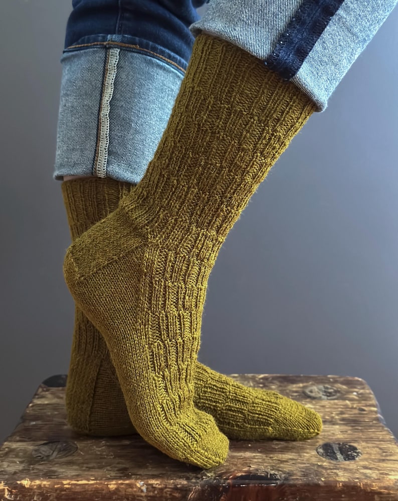 May include: A pair of olive green knitted socks with a textured pattern. The socks are worn with blue jeans. The socks are made of a soft, comfortable material.