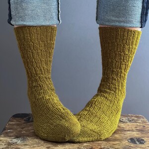 May include: Olive green knitted socks with a textured pattern, worn with blue jeans. The socks are on a wooden stool. The socks are a warm, earthy tone. The socks are approximately 30 cm in length.
