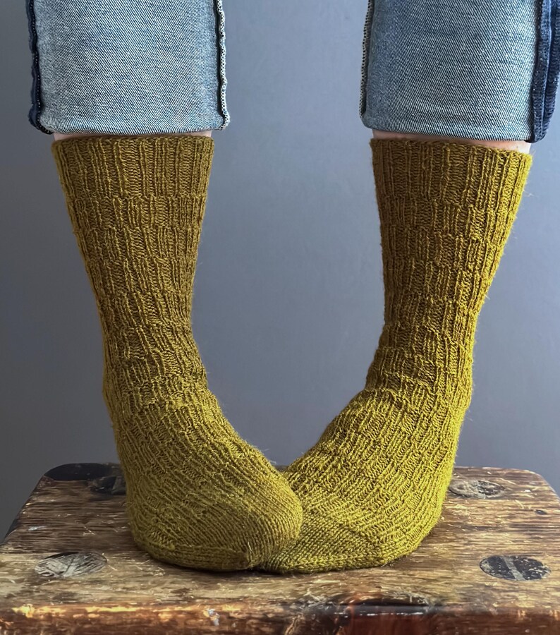 May include: Olive green knitted socks with a textured pattern, worn with blue jeans. The socks are on a wooden stool. The socks are a warm, earthy tone. The socks are approximately 30 cm in length.