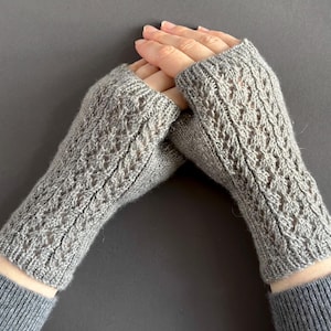 Könnte beinhalten: Ein Paar graue, gestrickte Fingerhandschuhe. Die Handschuhe haben ein strukturiertes, offenes Design und sind aus einem weichen, hellgrauen Garn gefertigt. Die Handschuhe werden an den Händen einer Person gezeigt, wobei die Finger frei sind.