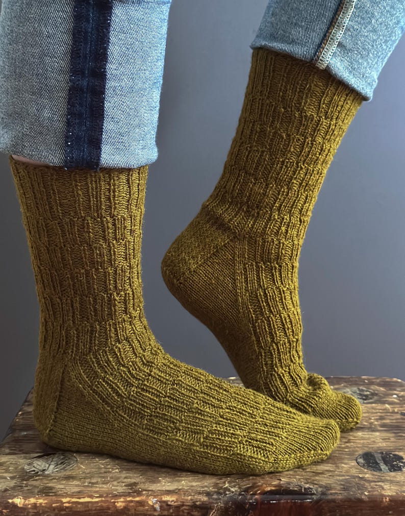 May include: A pair of olive green knitted socks with a textured pattern. The socks are worn with blue jeans. The socks are made of a soft, comfortable material.