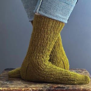 May include: Mustard-coloured knitted socks with a textured pattern. The socks are worn with blue jeans, and the image shows the socks from the ankles down. The socks are resting on a wooden surface.
