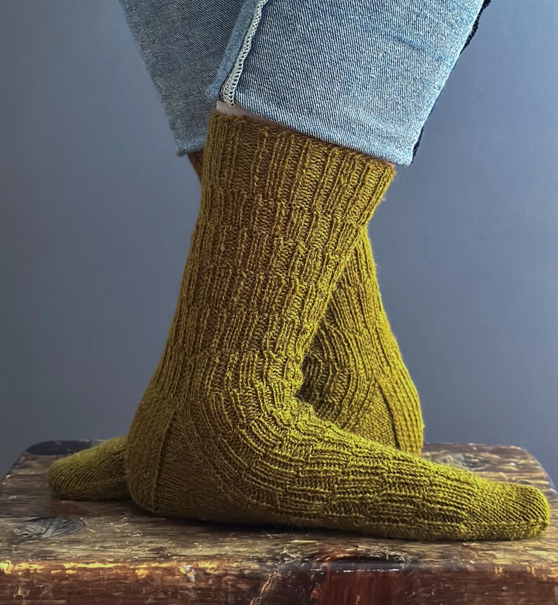 May include: Mustard-coloured knitted socks with a textured pattern. The socks are worn with blue jeans, and the image shows the socks from the ankles down. The socks are resting on a wooden surface.