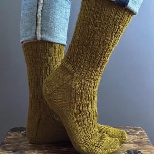May include: Close-up of mustard-coloured knitted socks with a textured pattern. The socks are worn with blue jeans. The socks are made of a soft, comfortable material.