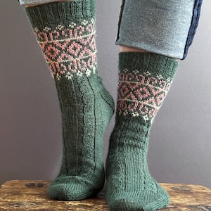 May include: A pair of mid-calf knit socks in a deep forest green colour. The socks feature a ribbed cuff, a decorative cable knit pattern running down the front, and a band of intricate geometric fair isle detailing in pink and cream tones.