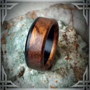 May include: A wooden ring with a black inlay. The ring is made of a dark wood with a natural grain pattern. The black inlay is a thin strip that runs around the entire circumference of the ring.
