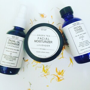 Puede incluir: Tres productos para el cuidado de la piel: un limpiador facial en aceite, una crema hidratante facial de té verde y un tónico facial. Los productos están en botellas azul oscuro y un tarro negro, con etiquetas blancas. Pétalos de naranja secos están esparcidos alrededor.
