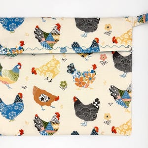 May include: A rectangular cream-colored fabric pouch featuring a repeating pattern of colorful chickens. The chickens have various patterns and colors, including blue, orange, and black. The pouch has a small loop on the side.