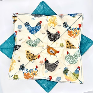 May include: A white fabric pot holder with a blue, green, and yellow floral pattern featuring chickens. The pot holder is quilted and has a teal blue fabric backing.
