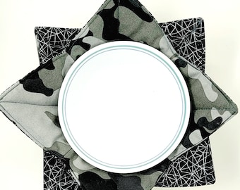 May include: A white ceramic plate sits on top of two fabric pot holders. The top pot holder has a camouflage pattern in shades of green and black. The bottom pot holder has a black and white geometric pattern.