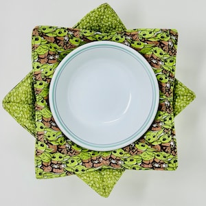 May include: A set of two green and brown fabric bowl cozies featuring a repeating pattern of a popular fictional character. The cozies are folded and a white bowl is sitting on top of them.