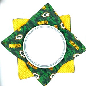 May include: Green and yellow fabric pot holder with a green camouflage pattern and the word "Packers" printed on it. The pot holder is folded in half and a white plate is sitting on top of it.