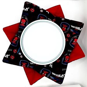 May include: A white plate with a teal rim sits atop a star-shaped trivet. The trivet has a red base and a black top patterned with Spider-Man graphics. The design features the superhero's face and the words "Spider-Man".