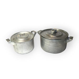 May include: Two silver-colored metal cooking pots with matching lids and side handles. The pots have a matte finish and a cylindrical shape. One pot is smaller than the other, and both feature a simple, functional design for kitchen use.