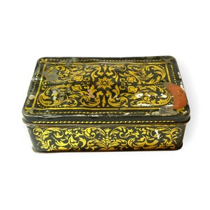 May include: A rectangular metal storage tin featuring a black base with intricate gold filigree patterns covering the lid and sides. The surface shows signs of wear, including small areas of paint loss and a small orange sticker on the lid.