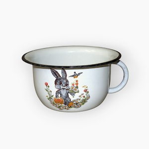 Könnte beinhalten: Eine weiße Emailletasse mit schwarzem Rand und einem Comic-Hasen, der Blumen und einen Korb hält. Der Hase ist von Blumen und einem Vogel umgeben.