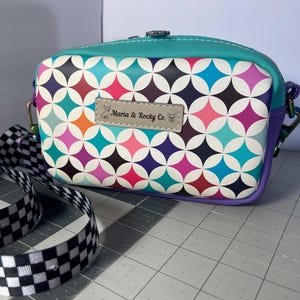 May include: A rectangular crossbody bag featuring a colorful geometric pattern of white, teal, pink, orange, and dark blue shapes. A rectangular label on the front reads Maria & Rocky Co. with a dog icon. It has a black and white checkered strap.