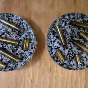 May include: Two round coasters with a speckled camouflage pattern, each embedded with several brass bullet casings. The coasters are set on a wooden surface, showcasing a unique, industrial design.