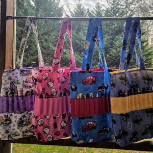 May include: Four colorful tote bags with different cartoon characters and designs. Each bag has a pocket for crayons or markers. The bags are hanging on a metal rod.
