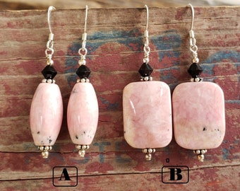 Pode incluir: Dois pares de brincos com pingentes de pedra rosa, ovais e retangulares, polidos. Cada brinco tem um gancho de prata, uma conta preta e detalhes prateados. Os brincos são exibidos em uma superfície de madeira rústica.