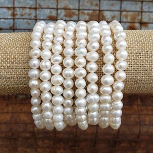May include: A stack of white pearl bracelets, each strand composed of round pearls, displayed on a beige fabric background.