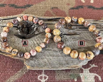 Pode incluir: Duas pulseiras feitas de pedras marrons, vermelhas e brancas. As pulseiras estão sobre uma superfície de madeira. As pulseiras são etiquetadas com A e B.