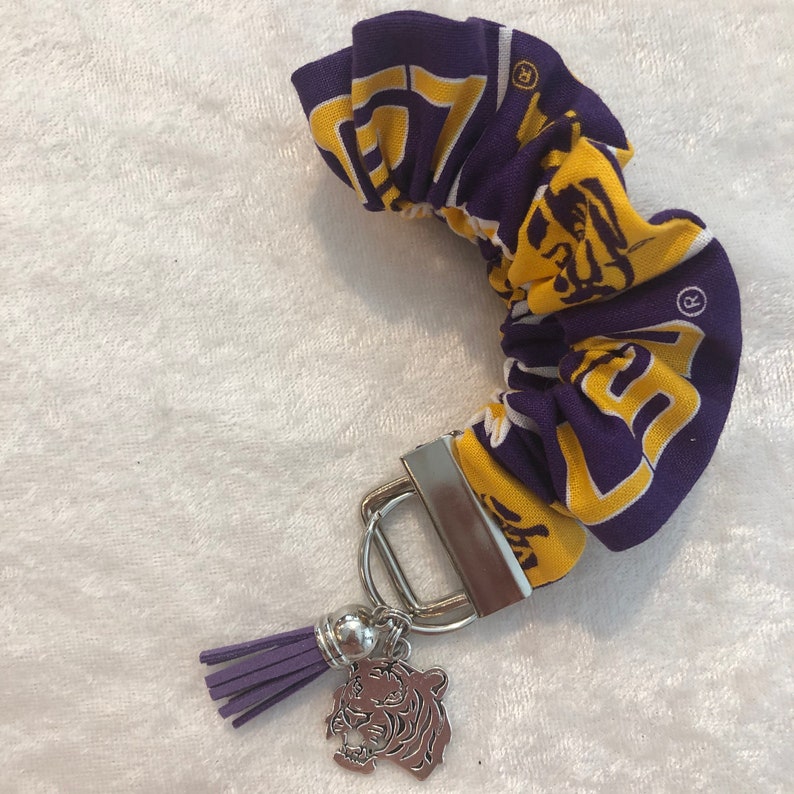 May include: Purple and gold fabric scrunchie with a tiger charm and a silver key ring. The scrunchie has the LSU logo on it.