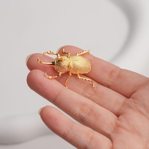 Puede incluir: Un broche de escarabajo de color dorado con detalles intrincados. El broche está hecho de metal y tiene un diseño realista.