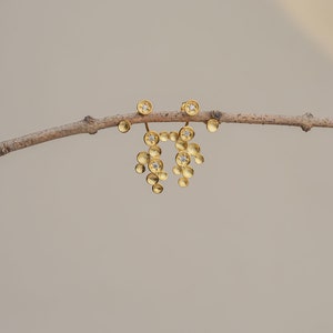 Puede incluir: Un par de pendientes de oro con pequeños colgantes redondos texturizados. Cada colgante tiene una pequeña piedra redonda transparente en el centro.