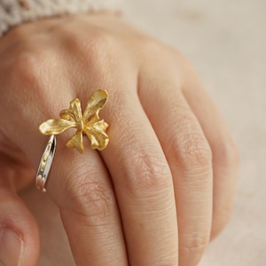 Puede incluir: Un anillo de oro con un diseño detallado de una flor de orquídea. El anillo se lleva en un dedo.