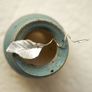 May include: A silver leaf charm with a twisted wire stem, resting on a blue ceramic surface.