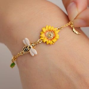 Puede incluir: Pulsera dorada con un dije de girasol y abeja. El girasol tiene pétalos amarillos y un centro rosa, mientras que la abeja tiene alas blancas y rayas negras. También hay un pequeño dije de hoja.