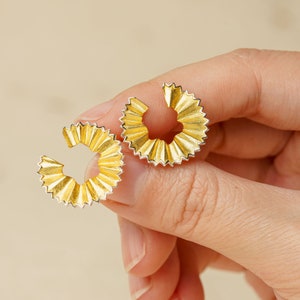 May include: A pair of gold-toned earrings with a unique, textured, fan-like design. The earrings are made of metal and have a circular shape.