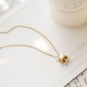 May include: A gold-colored necklace featuring a small, geometric elephant pendant. The pendant is attached to a delicate chain. The necklace is displayed on a white surface with text in the background.