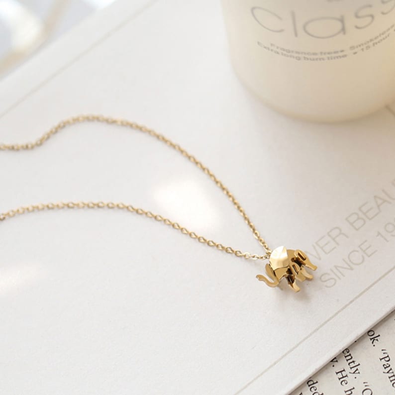 May include: A gold-colored necklace featuring a small, geometric elephant pendant. The pendant is attached to a delicate chain. The necklace is displayed on a white surface with text in the background.