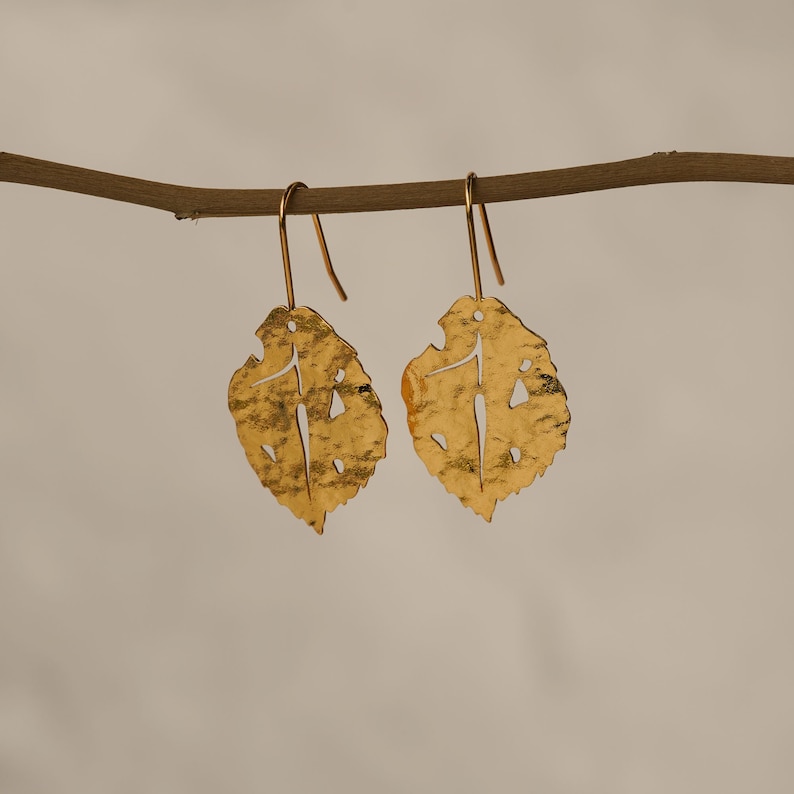 May include: A pair of gold leaf-shaped earrings with a delicate, cutout design. The earrings hang from a simple gold hook.