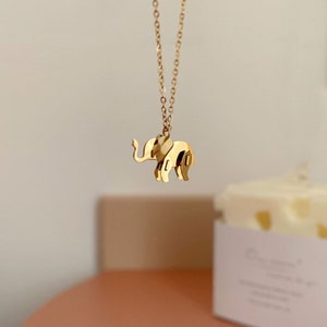 May include: A gold-colored elephant pendant necklace. The pendant is a stylized, three-dimensional elephant with detailed features. The necklace chain is delicate and also gold-colored. The background is a soft, neutral color.