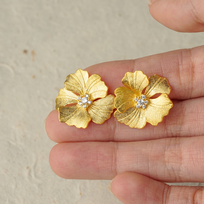 May include: A pair of gold flower-shaped earrings with a small clear crystal in the center of each flower.