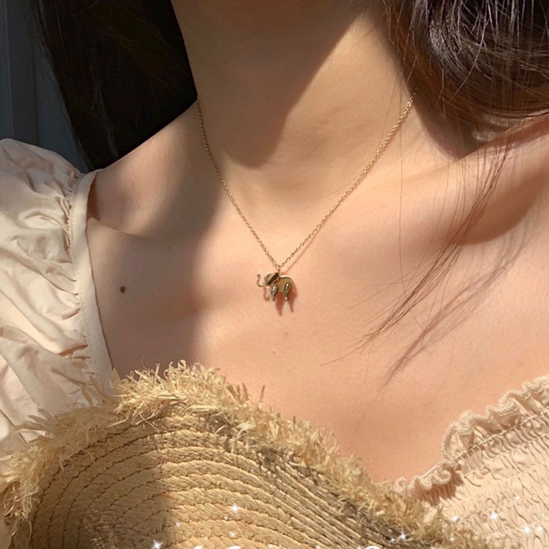 May include: A gold-colored necklace with a small elephant pendant. The elephant pendant has detailed features and is suspended from a delicate chain. The necklace is worn against a light-colored top, creating a contrast.