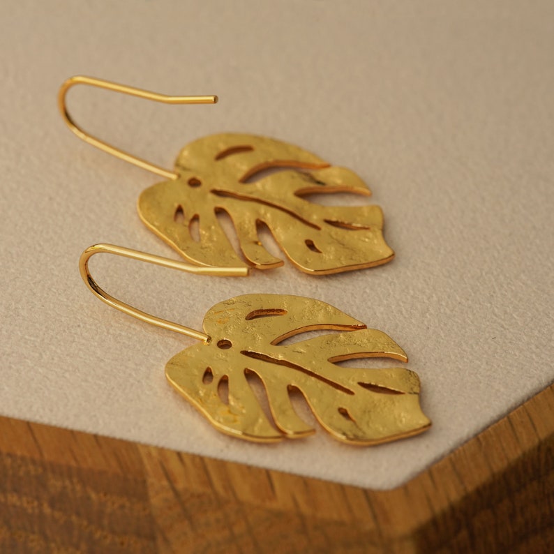 May include: A pair of gold-tone earrings shaped like monstera leaves. The leaves have a textured surface and are slightly curved.